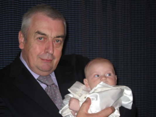 Alan Wilkinson, smartly dressed in dark suit and grey tie, cradles a baby all in white.