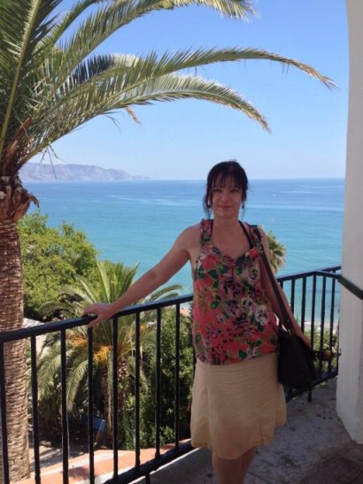 Holiday photo of Anne Bitton, from a balcony overlooking the sea, framed by a palm tree.