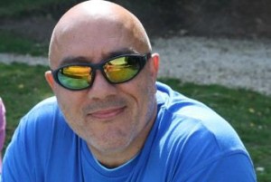 Photo of Andy Kemp sitting outdoors looking relaxed wearing reflective sunglasses and a blue t-shirt.