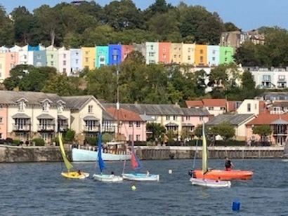 A harbour with small sailboats in the foreground and brightly painted houses on a hill in the background