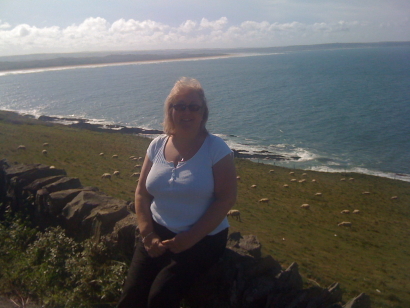 Carole, perched on a coastal wall. Behind her ia grassy bank on which sheep graze which leads down to the sea 