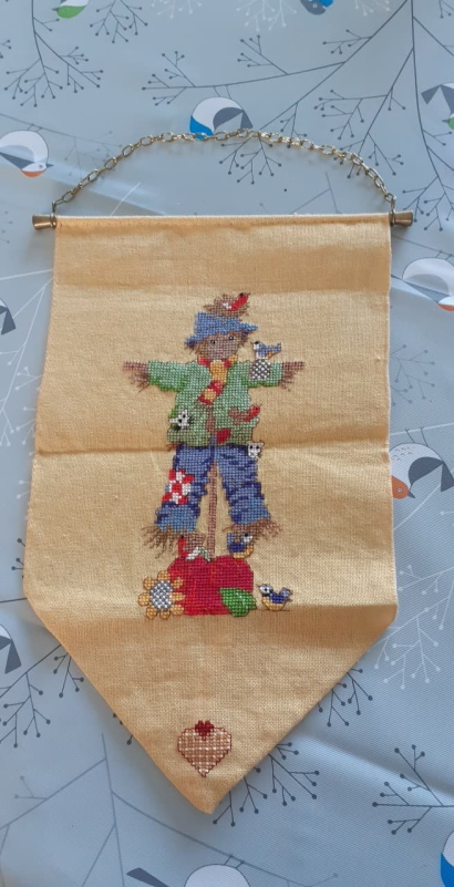 Cross-stitched scarecrow onto a pennant, Scarecrow has blue hat, red an white scarf, green shirt and blue trousers with a red and white checked patch. A couple of birds are perching on it.