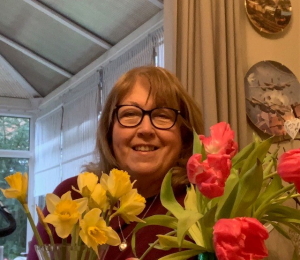 Carole, indoors, smiling behind a display of red and yellow flowers