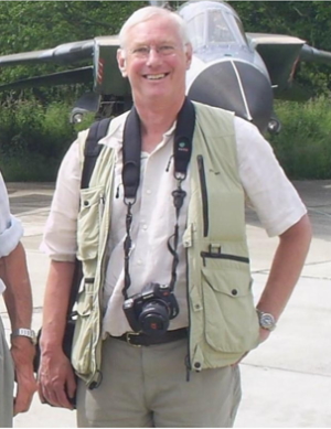 John smiling, standing in from of an aircraft, camera slung around his neck.