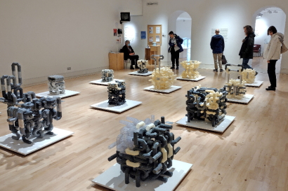 Black and cream coloured cubic sculptures of the interlocking letters on low grey plinths distributed over the wooden museum floor