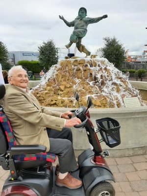 A smiling Frank sits on a mobility scooter in front of the 'Jolly Fisherman' fountain