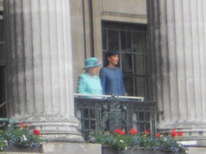 Queen Elizabeth and the then Duchess of Cambridge stand on a balcony. Both are dressed in blue, the queen in a lighter shade
