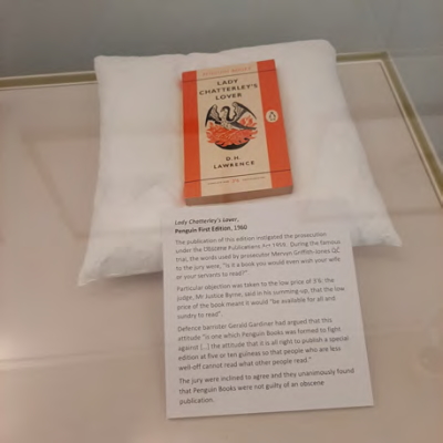 A first edition copy of 'Lady Chatterley's Lover' sits on a white cushion, with a note about the book.