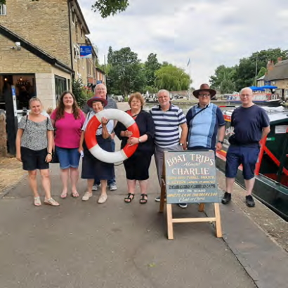 A group of eight pose on the edge of the canal in front of the boat trip sign. Two are holding a large lifebuoy ring